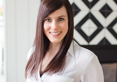 Woman with long dark brown hair wearing a white blouse, smiling indoors with a black-and-white geometric wall in the background.